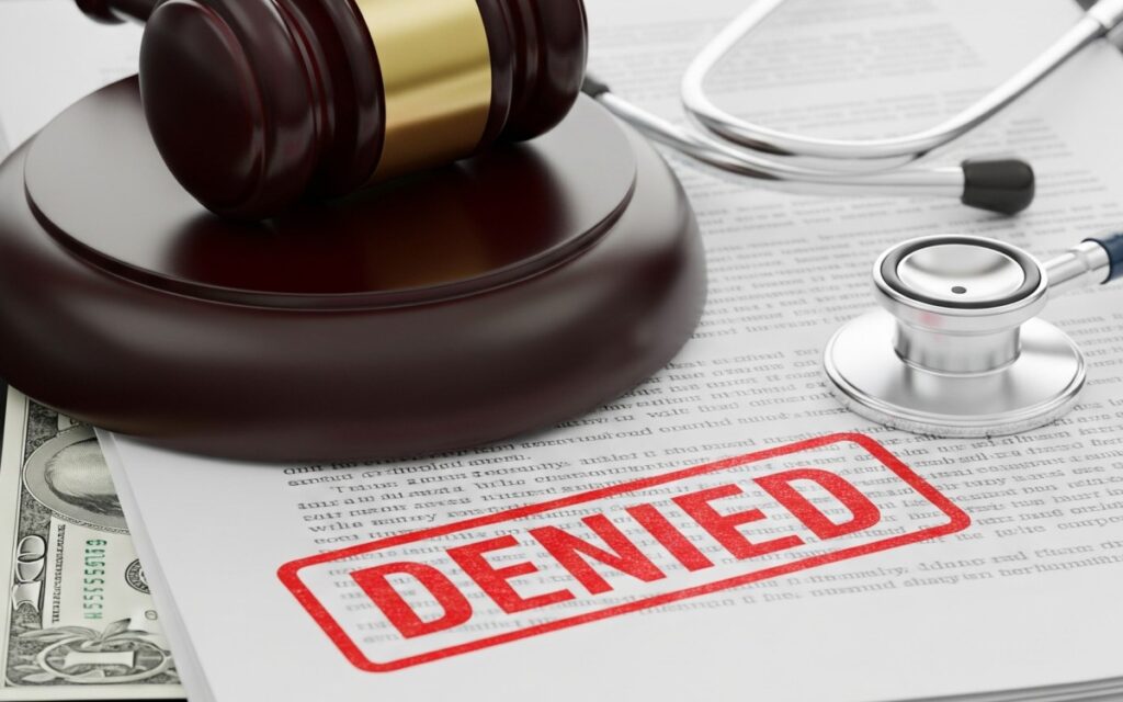 Conceptual image of a medical stethoscope, a legal gavel, and health insurance paperwork stamped with a red 'DENIED' mark, representing hospital revenue cycle strain.