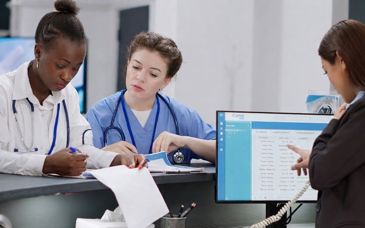 18e53a06-7368-47a6-ad42-f17a5b0c769a-2026-07-09 Two clinicians and an administrator reviewing claim denials at a hospital desk with digital records on screen