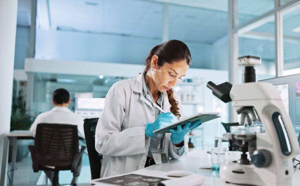 Scientist reviewing data on a tablet in a medical research laboratory with microscope and monitor